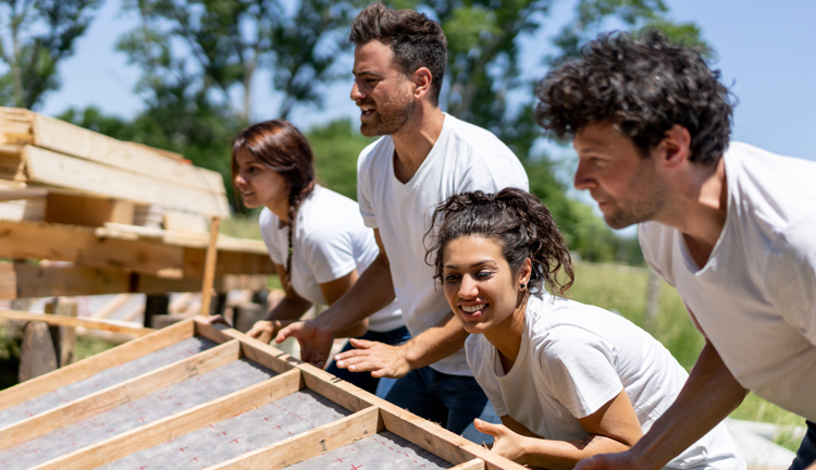 Four people all wearing white shirts outside are working together to lift up a wooden frame.